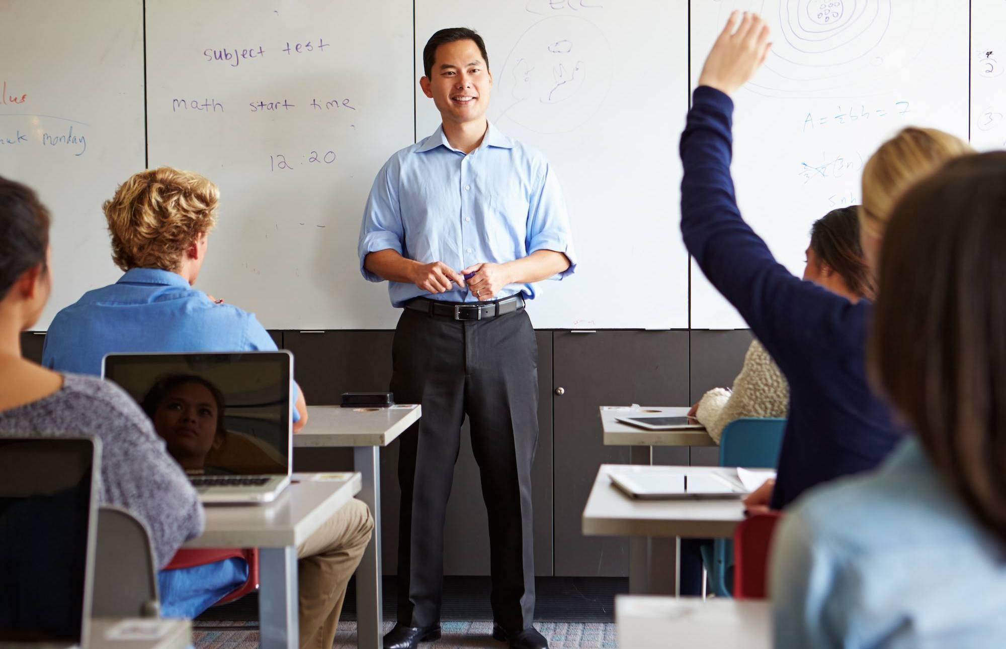 A high school math teacher stands in front of his class, smiling at a student raising her hand.