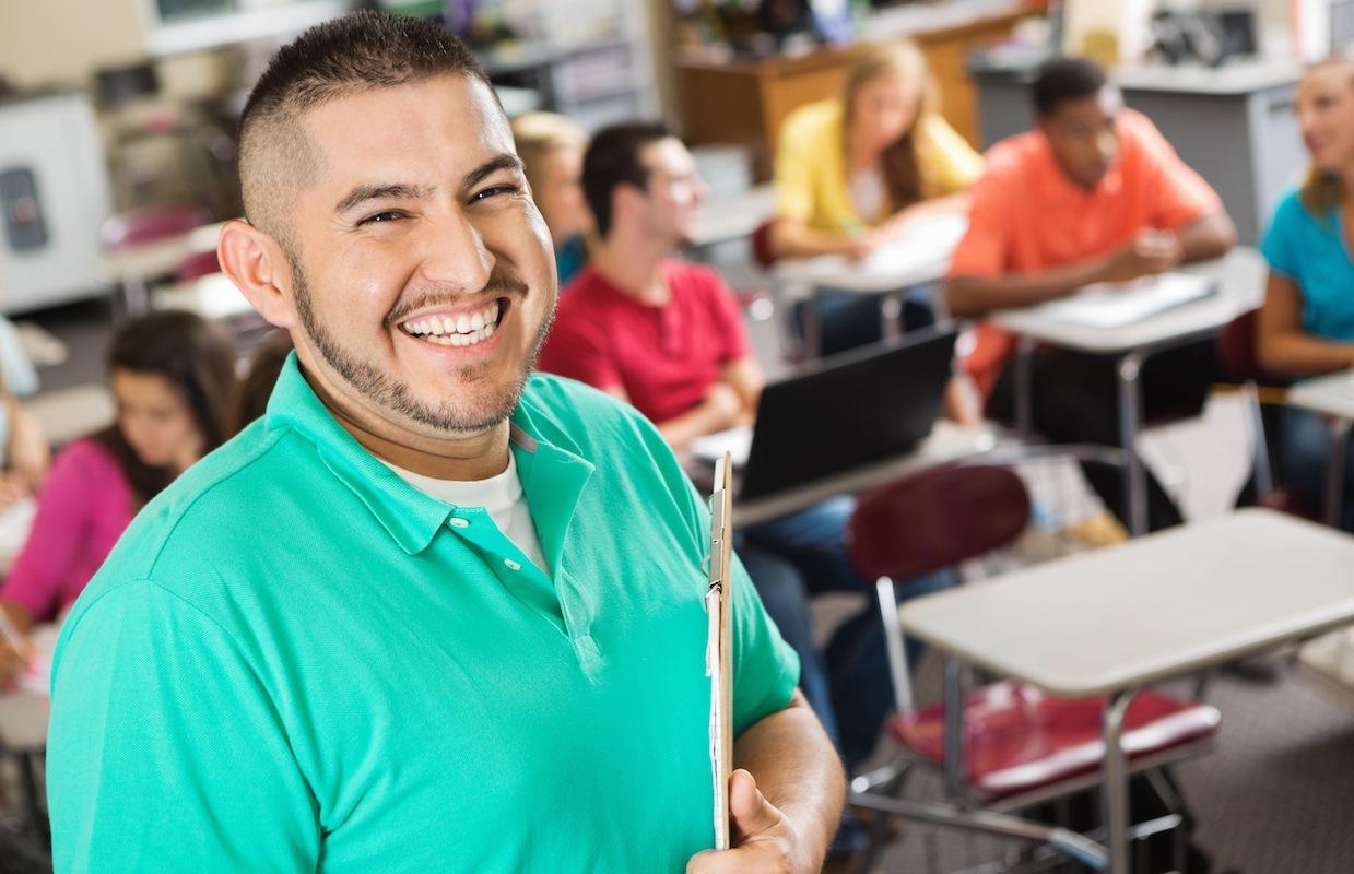 A high school teacher with a huge grin, standing in front of his class. He wears a sea green shirt and is holding a clipboard, smiling at the camera.