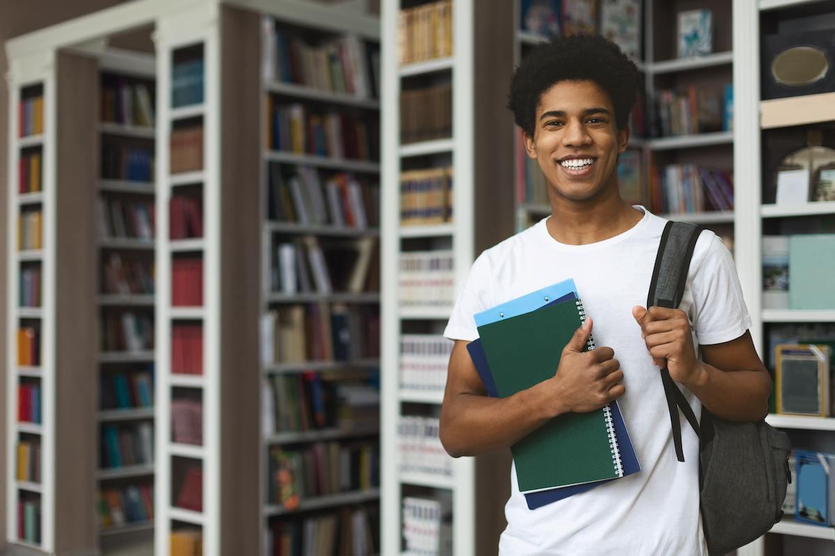 A future teacher standing in his campus library. He’s wearing a white T-shirt and holding books in one hand and a backpack on his shoulder with the other. 